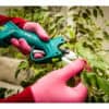 Mit rosa Handschuhen verwendet jemand eine grüne 16-V-Li-Ionen-Akku-Gartenschere, um belaubte Zweige im Innenbereich zu stutzen.