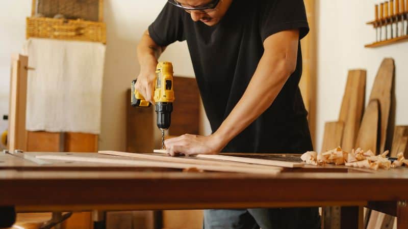 A person uses a yellow drill on a wooden plank, surrounded by wood shavings and tools in a workshop.
