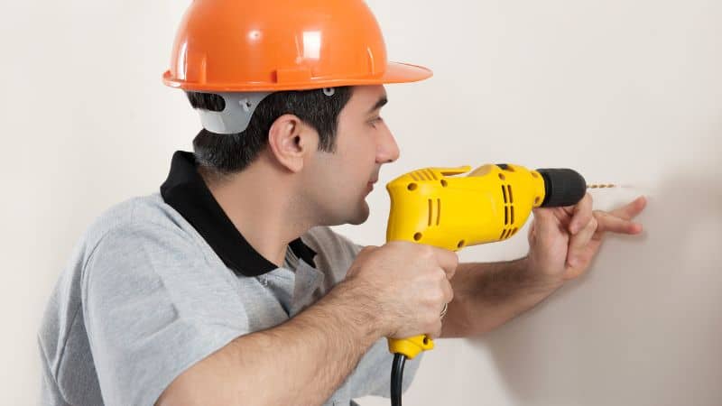 Worker in an orange hard hat using a yellow drill on a wall.
