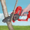 Someone prunes a tree branch with a red and black handheld chain saw in a grassy area under a blue sky.