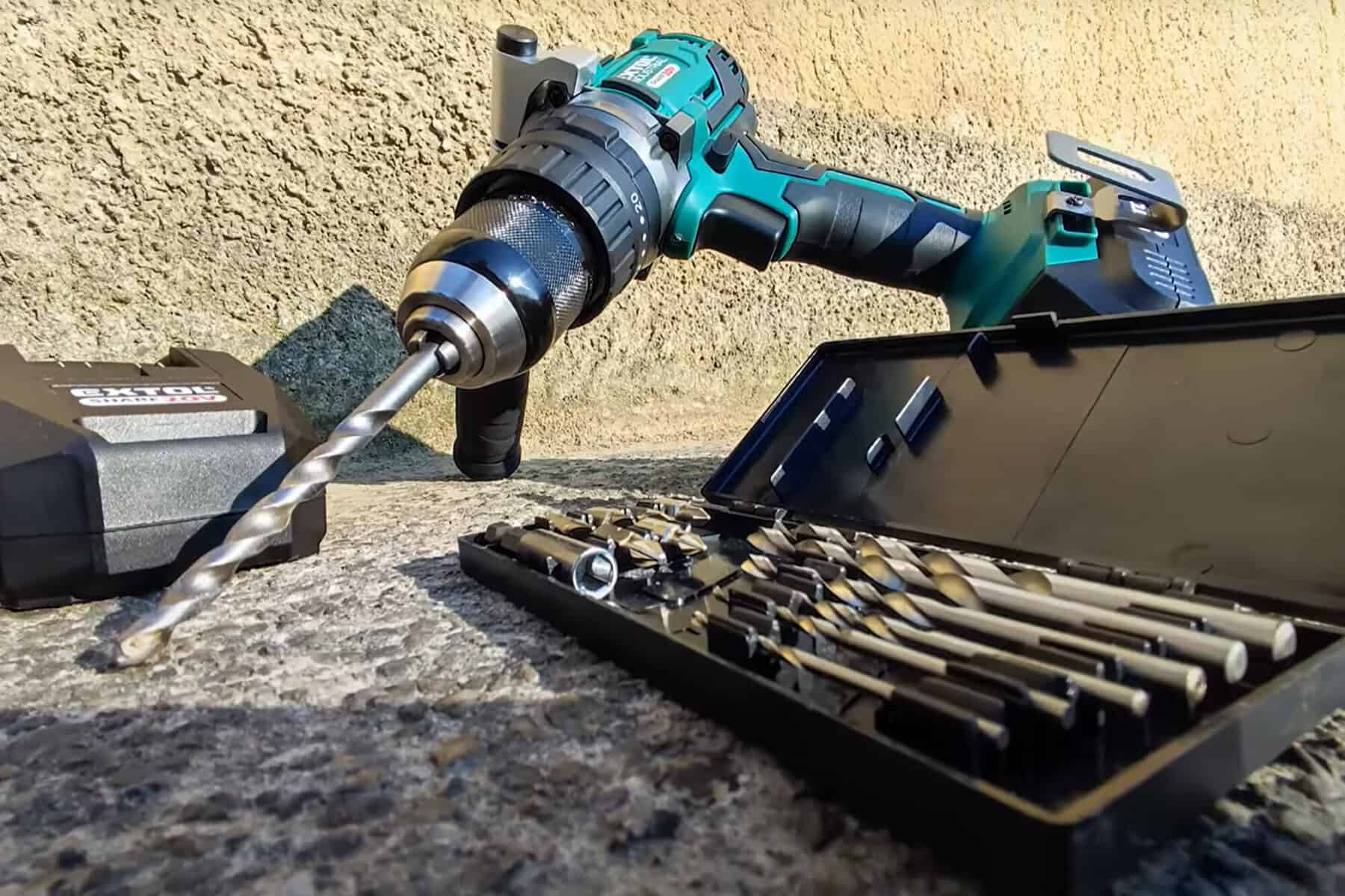 A cordless drill with a bit attached rests on a textured surface beside an open case of assorted drill bits.