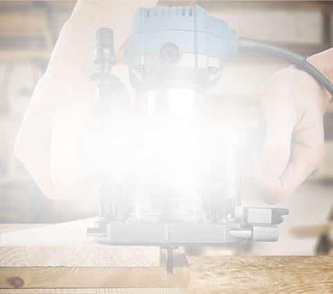 Someone shapes a wooden board’s edge with a handheld router in a workshop.