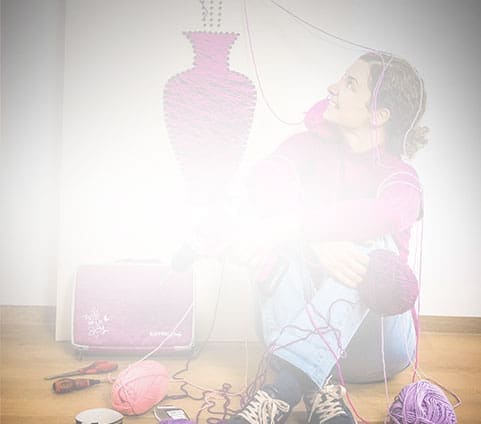 A person sits on the floor amid pink yarn, gazing at a half-finished vase-shaped yarn art on the wall.