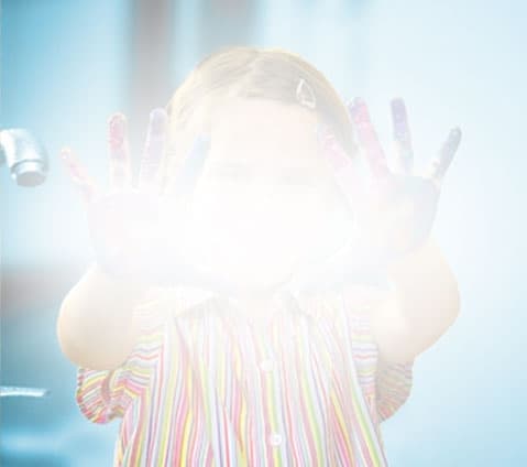 A child in stripes holds up painted hands, their face and palms obscured by bright light.