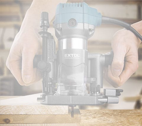 A person uses a handheld router to shape the edge of a wooden board in a workshop.