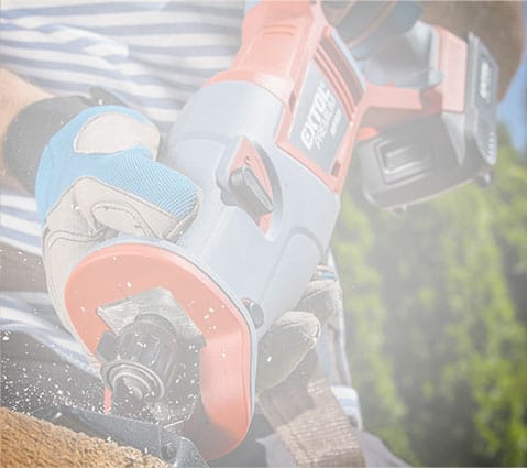 Wearing gloves, a person uses a cordless power tool to cut wood as sawdust flies.