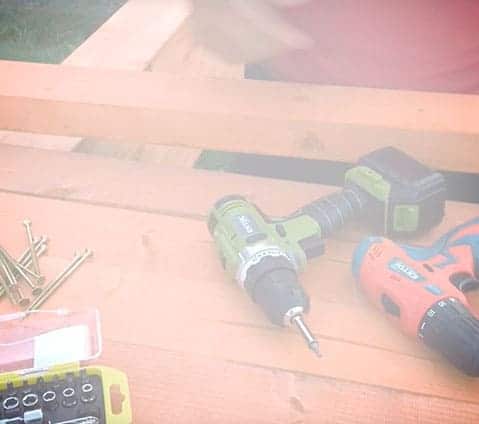 Two cordless drills, drill bits, and screws are on a wooden surface; a person is partly visible in the background.