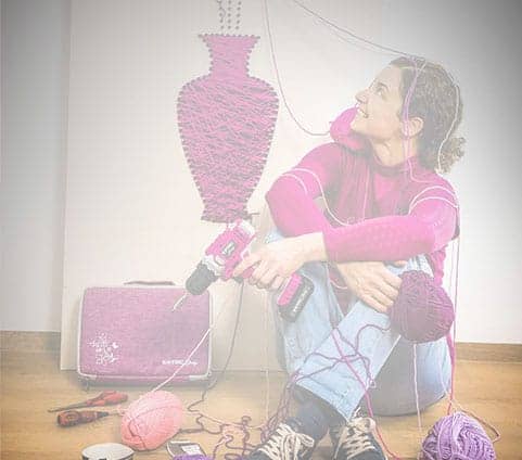 A person sits on the floor with a pink yarn-wrapped drill, yarn balls, and a yarn-covered vase hanging nearby.