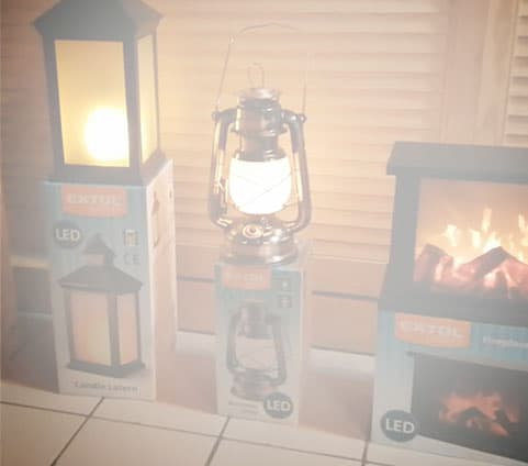 Three boxed LED lantern and fireplace products are displayed on a tiled floor before wooden blinds.