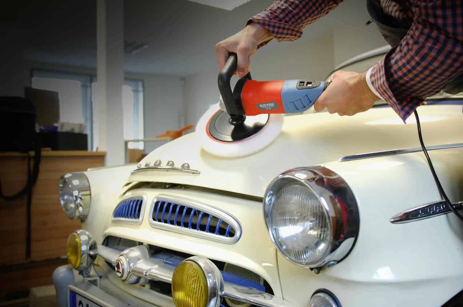 Someone uses an electric polisher on the hood of a vintage cream-colored car indoors.