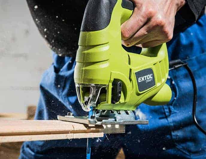 Someone cuts a wooden board with a green Extol Craft jigsaw, creating sawdust.