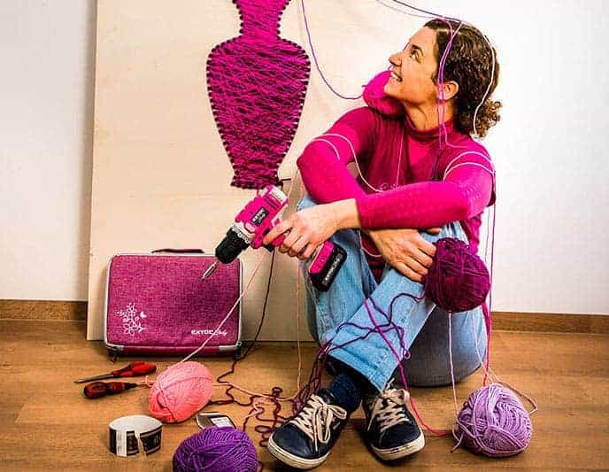 A woman uses yarn and a drill to make a pink and purple string art vase on canvas, surrounded by craft supplies.