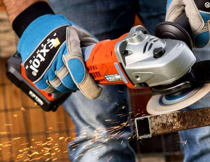 Gloved hands use an angle grinder to cut metal, sending visible sparks flying.