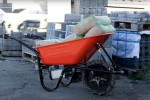 Red wheelbarrow with black frame holding cement bags at a construction site, surrounded by pallets of materials.