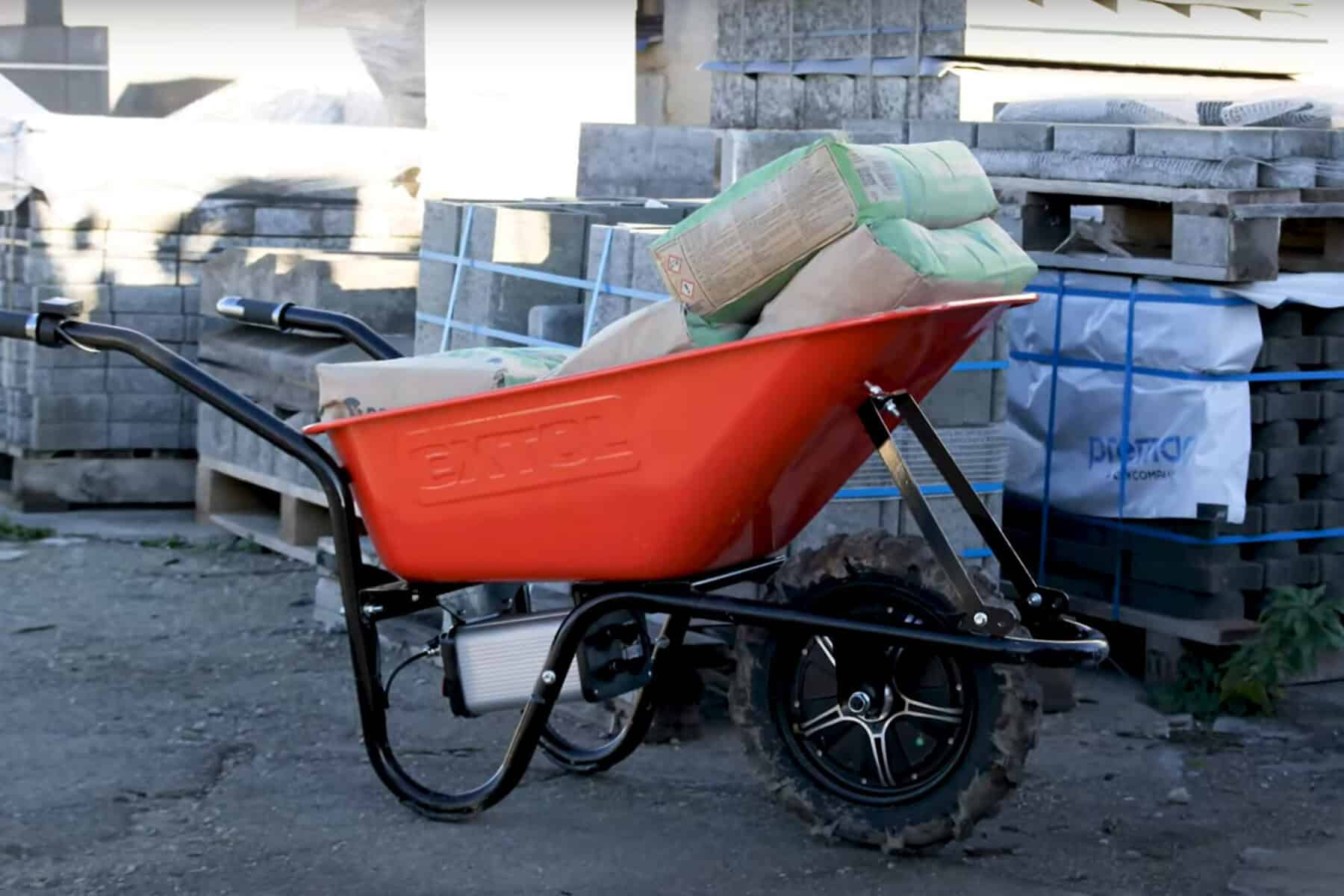 Red wheelbarrow with black frame holding cement bags at a construction site, surrounded by pallets of materials.