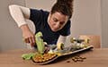 A woman hot glues snacks or nuts onto a skateboard on a table.