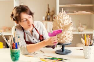 A woman uses a pink glue gun on a textured ceramic sculpture, near paintbrushes and art supplies on a table.