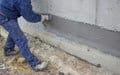 A worker in blue overalls uses a trowel to apply gray plaster to an exterior wall's base.