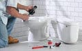 A person kneels by a toilet, using tools to repair it with plumbing supplies spread on the floor.