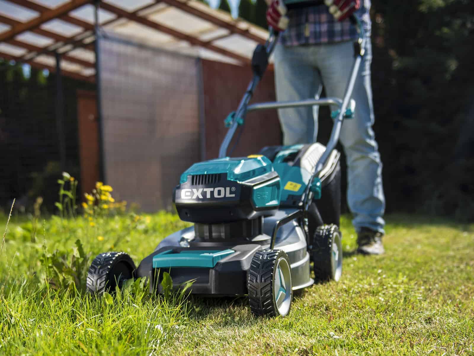 A person mows a grassy lawn with a teal-black Extol mower near a shed on a sunny day.