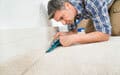Kneeling, a man uses a caulking gun to apply caulk along the base of a wall.