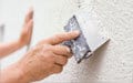 A person uses a putty knife to smooth plaster on a textured white wall.