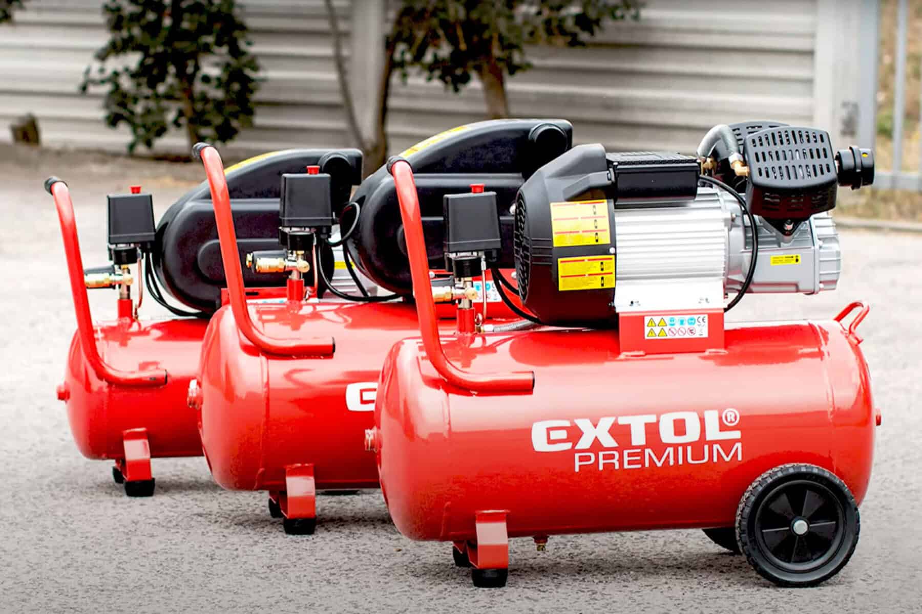 Three red Extol Premium air compressors in a row outdoors on pavement, with trees and a metal fence behind.