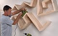 A man installs a geometric wooden shelf on a white wall using a power drill.