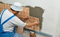 A worker in blue overalls and a white hat installs brown ceramic tiles on a wall with adhesive.