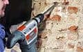 A person uses a power drill with a chisel to remove plaster or mortar from a brick wall.