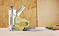 A green and silver staple gun sits on a wood surface with staples and a tool; wood paneling is behind.