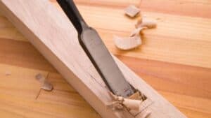 A wood chisel carves a groove into a light wooden board, with wood shavings and chips scattered around the cut.