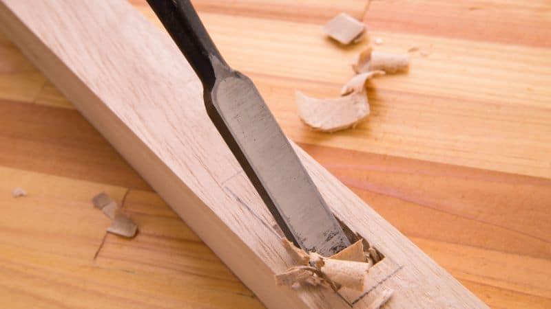 A wood chisel carves a groove into a light wooden board, with wood shavings and chips scattered around the cut.