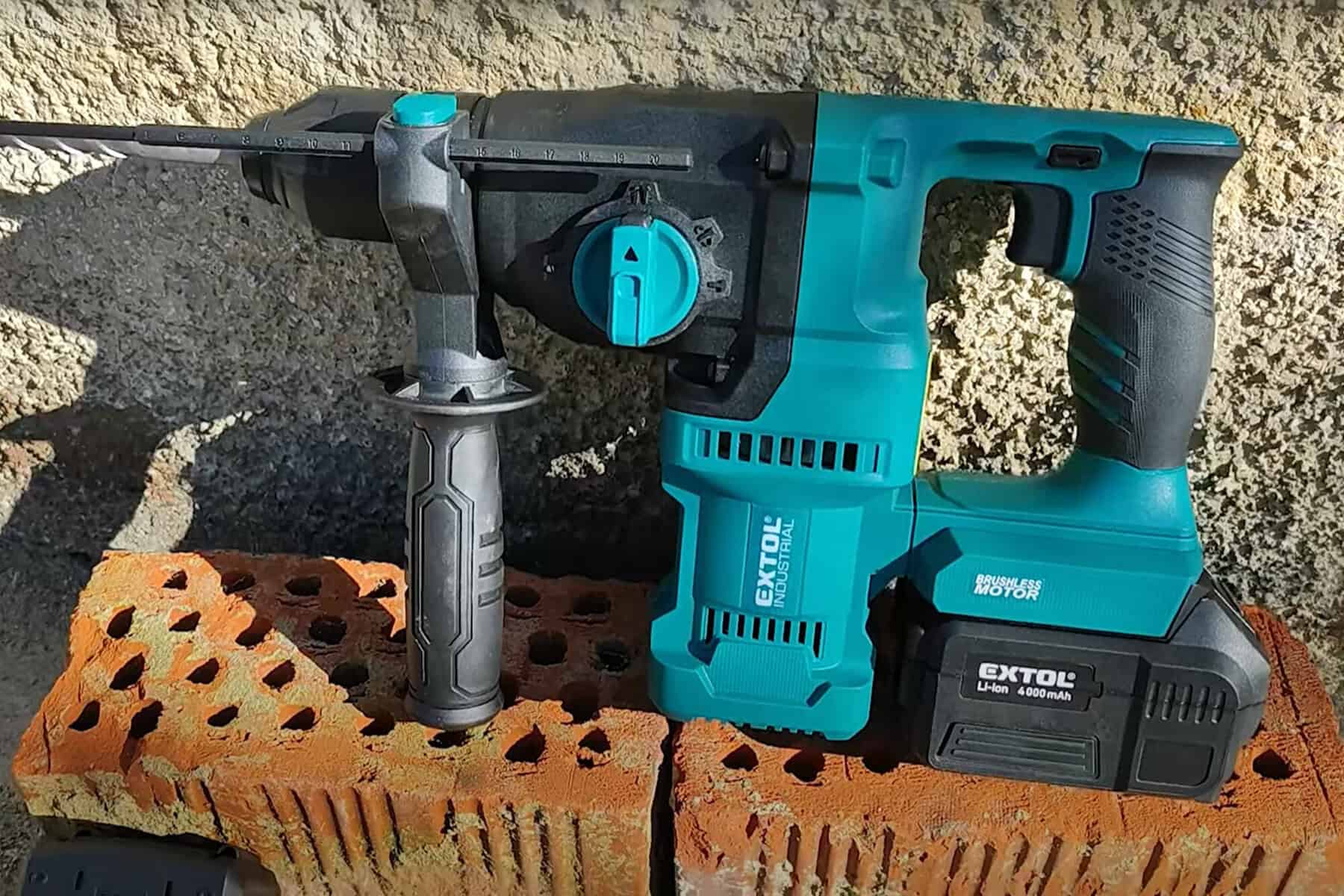 Cordless Extol rotary hammer drill with battery on perforated red bricks, set against a stone wall.
