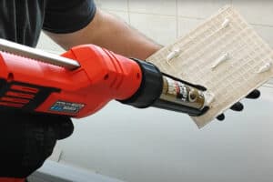 Someone uses a red electric caulking gun to apply adhesive to the back of a plastic panel.