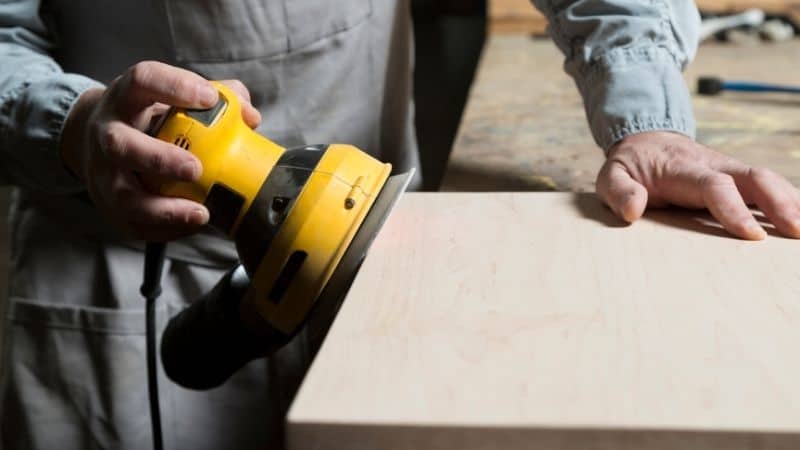A person sands a wooden board with a yellow electric sander on a workbench.