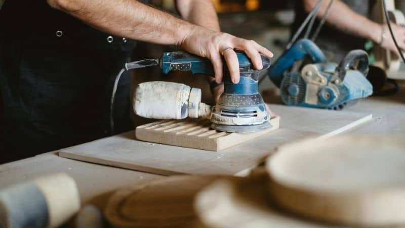 A person uses an electric sander on a wooden board at a workbench, with woodworking tools in the background.