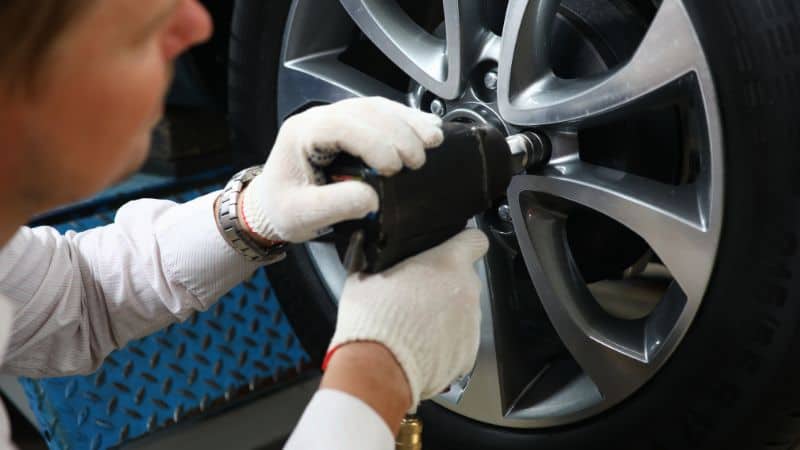 A person in white gloves uses a power tool on bolts of a car wheel.
