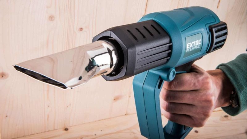 A person aims a blue Extol Industrial heat gun with wide nozzle at a light wooden surface.